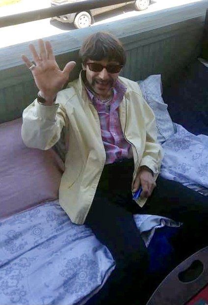 A man wearing sunglasses is sitting on a bed waving his hand.
