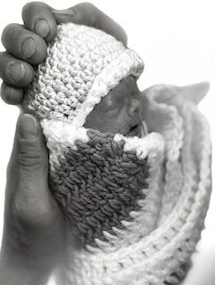 A person is holding a baby wrapped in a knitted scarf and hat.