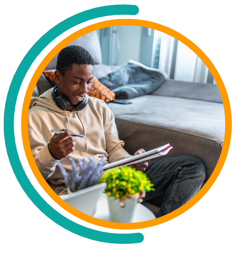Man sitting on floor, writing in notebook near couch, headphones on, smiling.