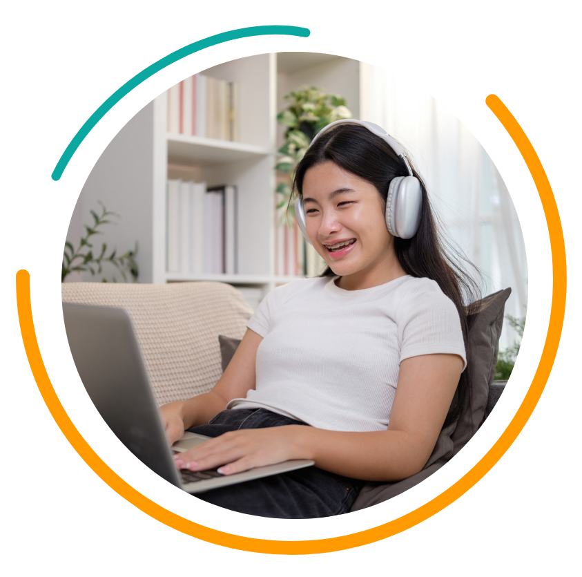 Teenage girl with headphones using a laptop, smiling. Sitting on a couch indoors, surrounded by books.