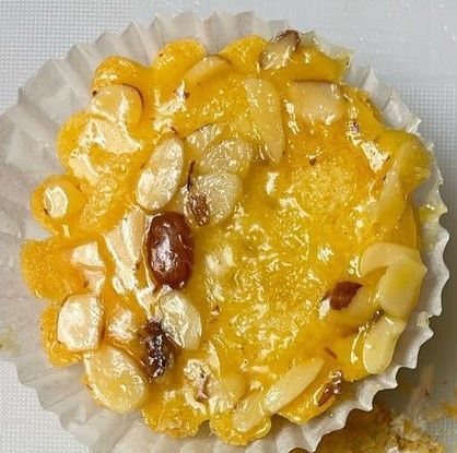 Small, yellow tart with almonds and a glazed topping, inside a white paper cup.