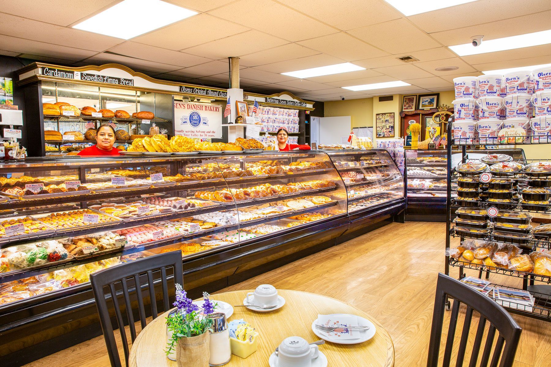 Inside a bakery, filled with pastries. Two people behind the counter. Round table with chairs in foreground.