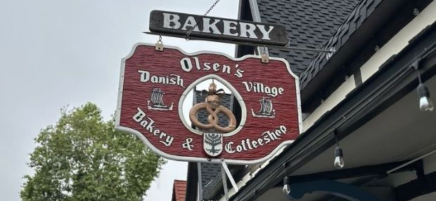 Inside a bakery, filled with pastries. Two people behind the counter. Round table with chairs in foreground.