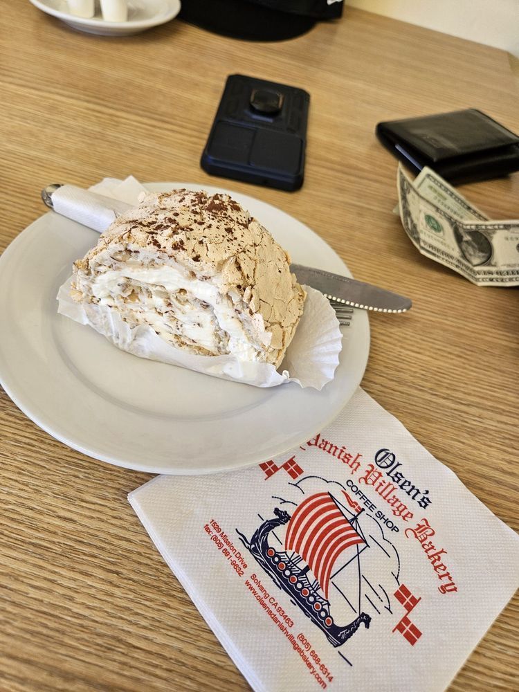 Danish pastry on a plate with a napkin from Olsen's Danish Village Bakery, money, and a phone on a table.