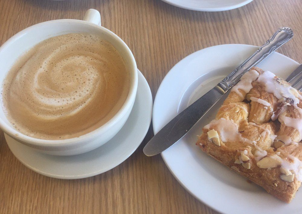 Cup of coffee with swirl pattern next to pastry on a plate with knife, on a wooden table.