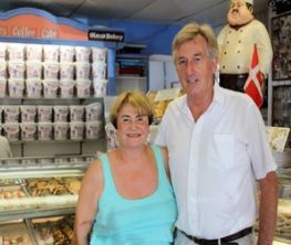 Couple standing in a bakery. Display case with pastries, shelves of containers. Chef statue with a flag in background.
