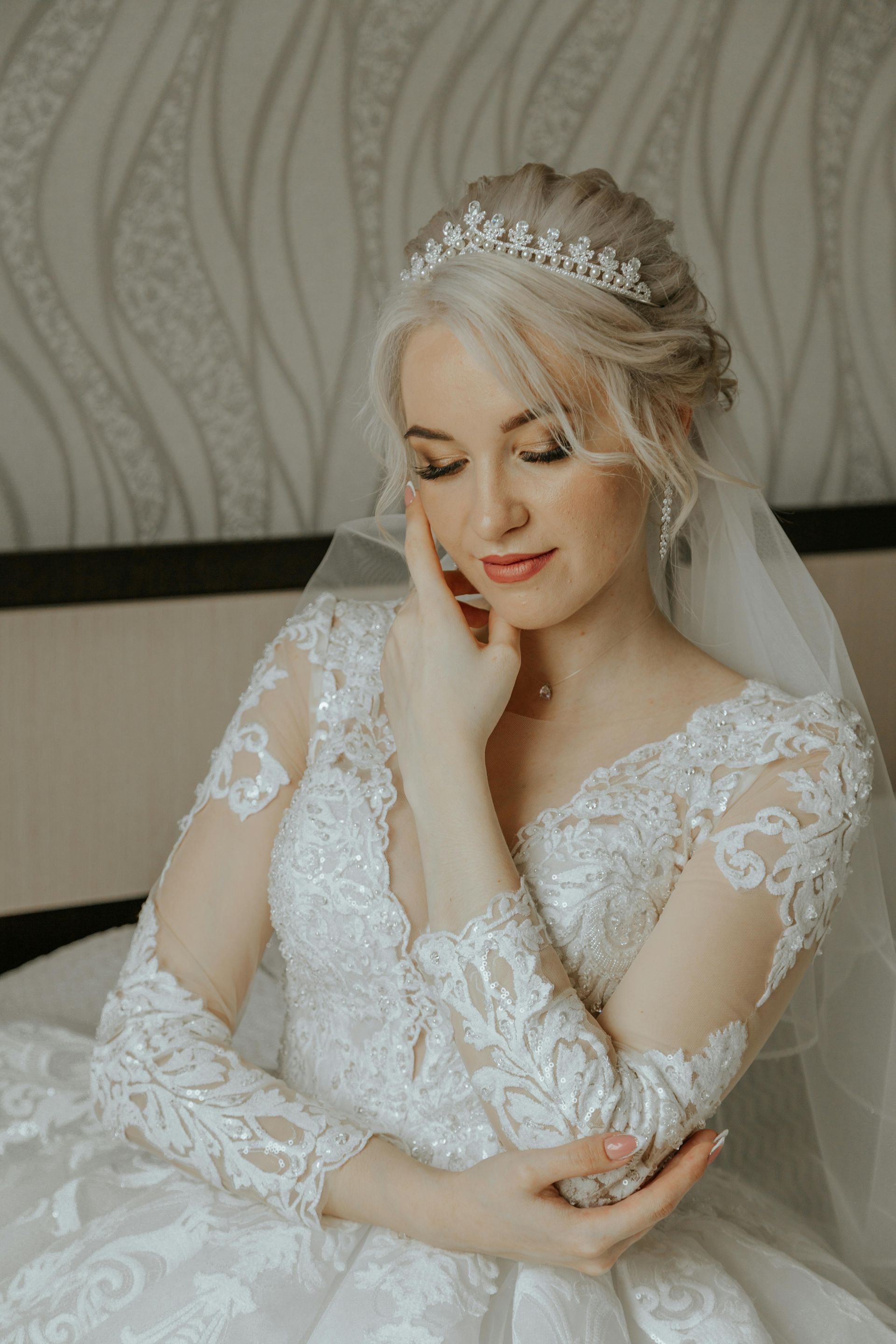 Bride in a white lace gown, tiara, veil, and updo, seated on a bed, touching her face with a serene expression.