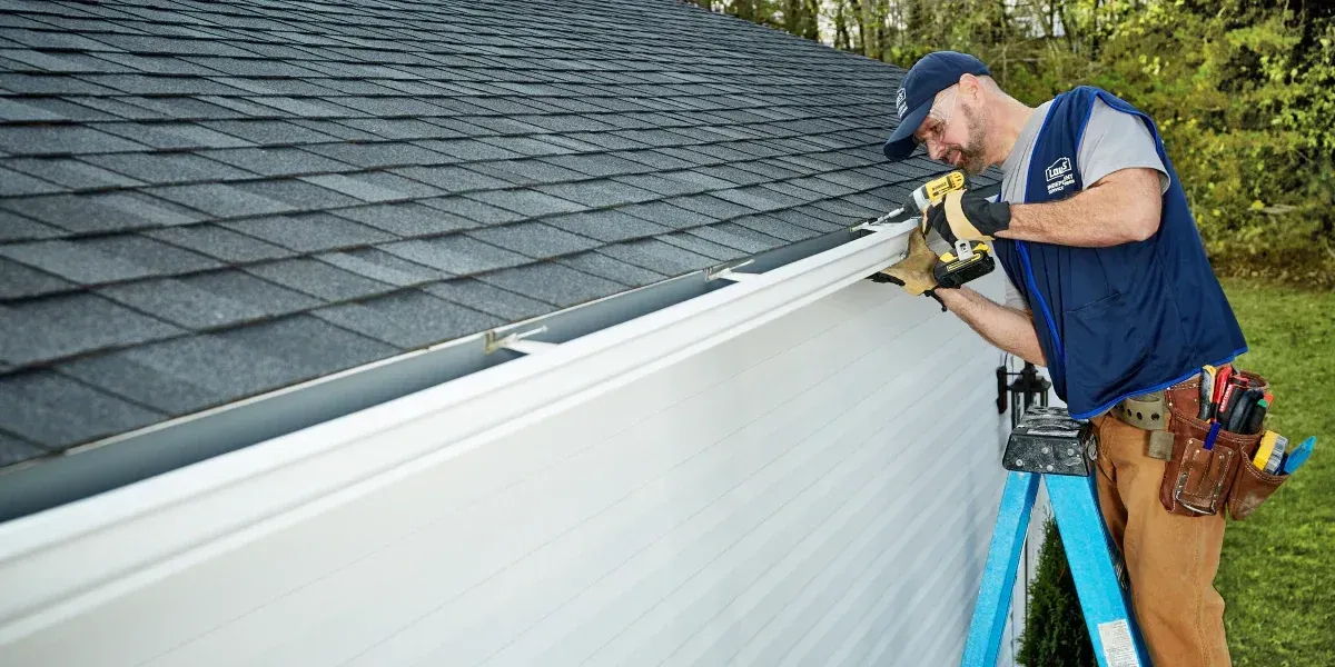 A worker on a ladder installing a gutter on a house's roof.