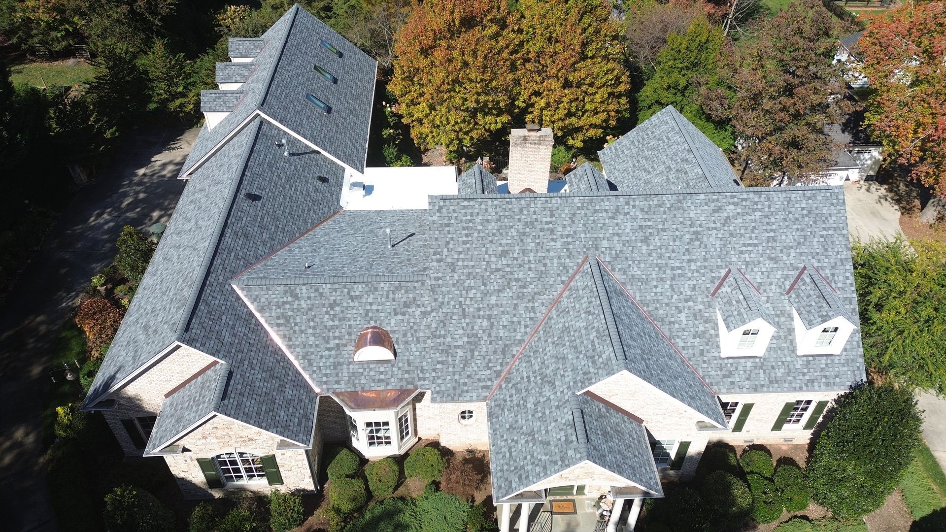 Aerial view of a large, multi-gabled house with a dark gray roof, surrounded by green grass and trees.