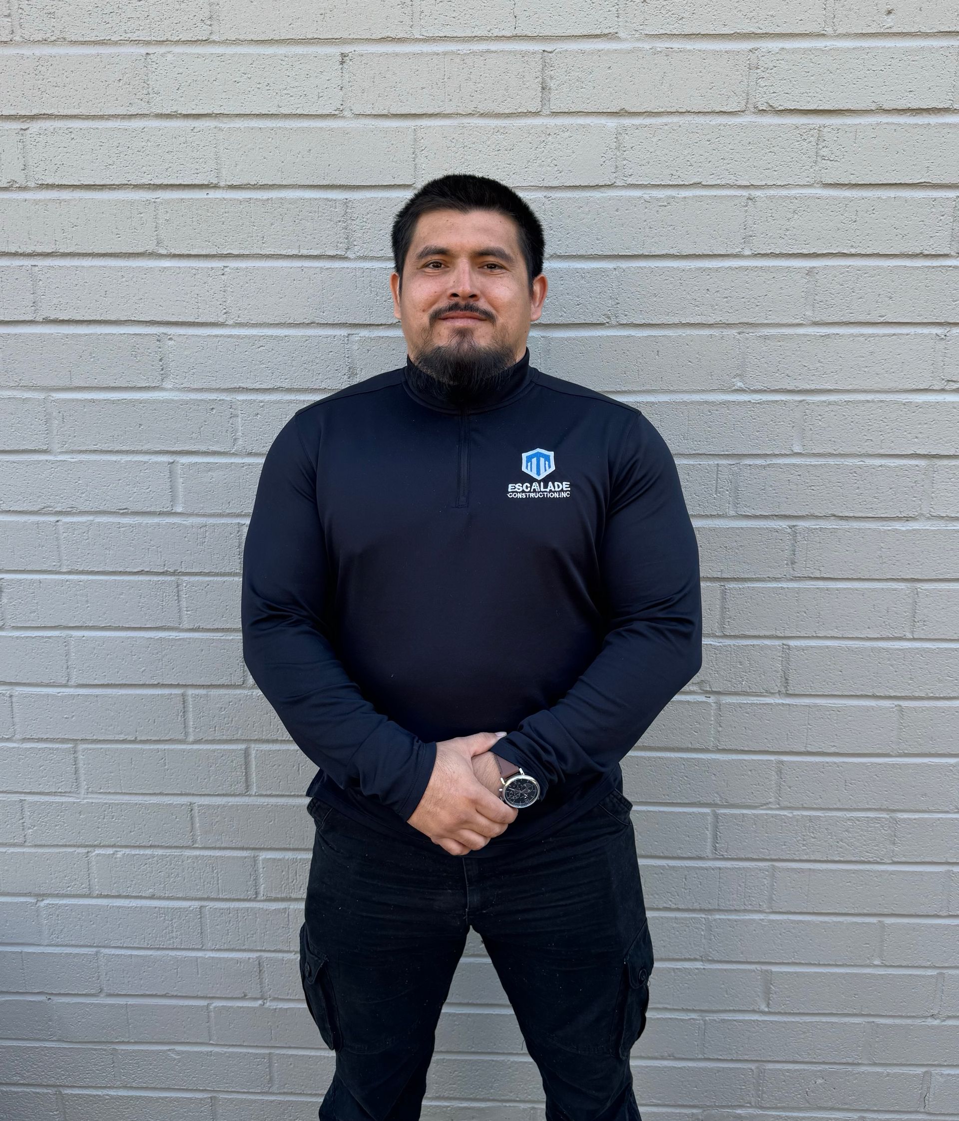 Man in black shirt and hat with arms crossed in front of a brick wall.