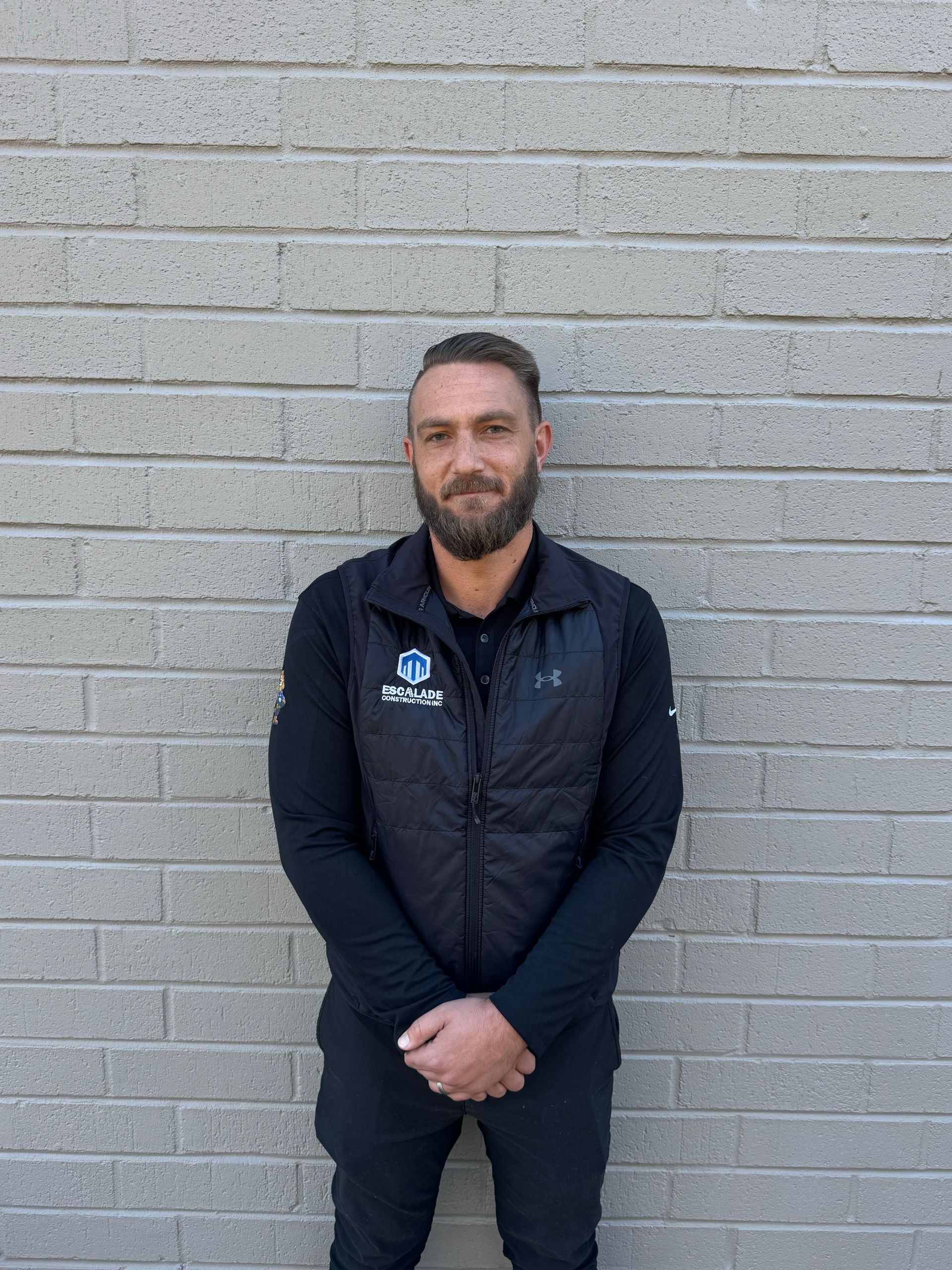 Man with a beard, wearing a "ROOF" hat and black polo, crosses arms, standing in front of a brick wall.