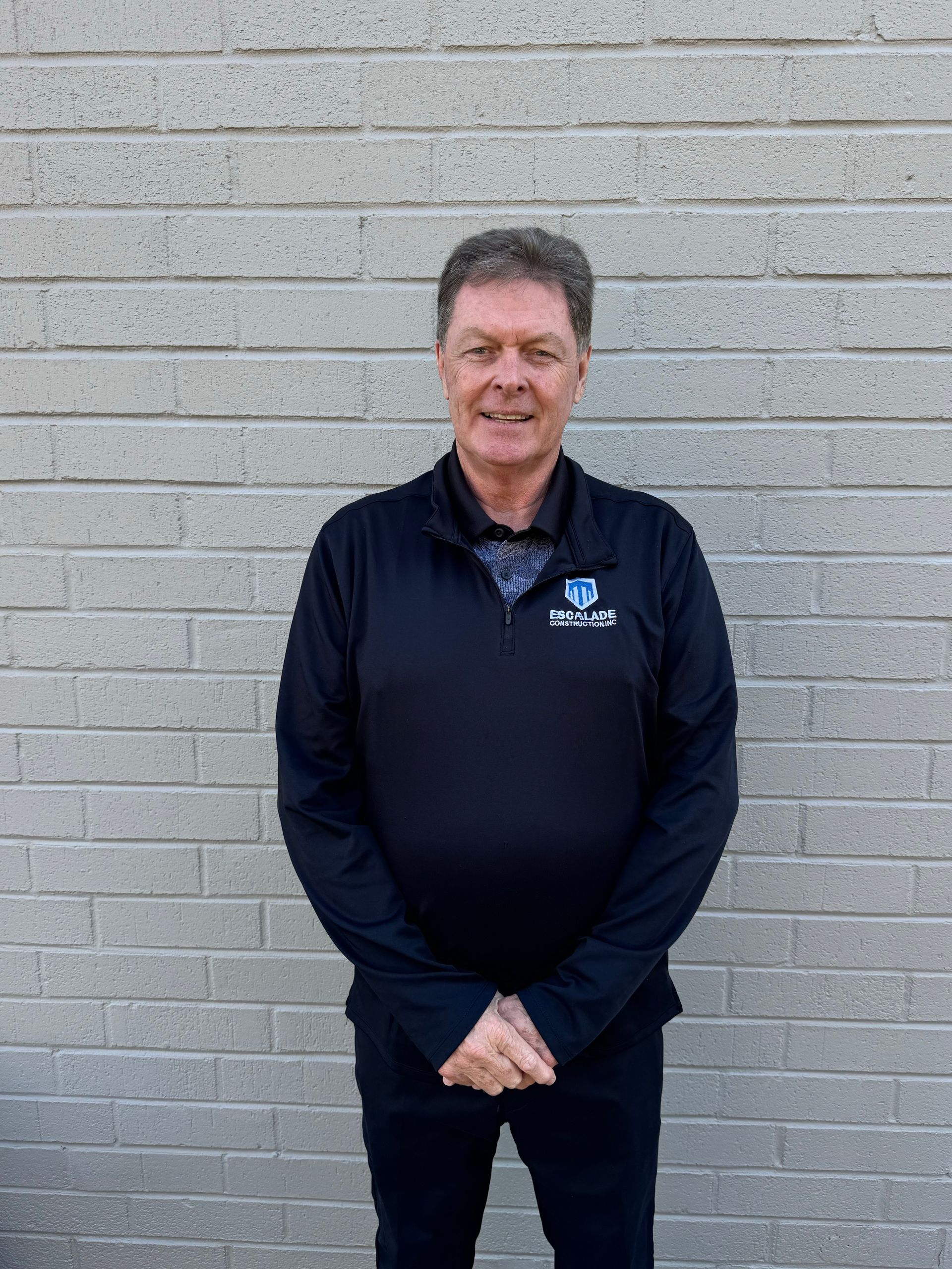 Man standing in front of a brick wall, wearing a black jacket and pants, smiling.