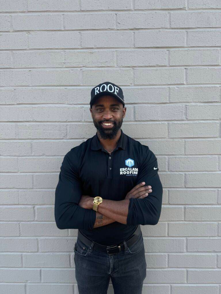 Man in black shirt and hat with arms crossed in front of a brick wall.