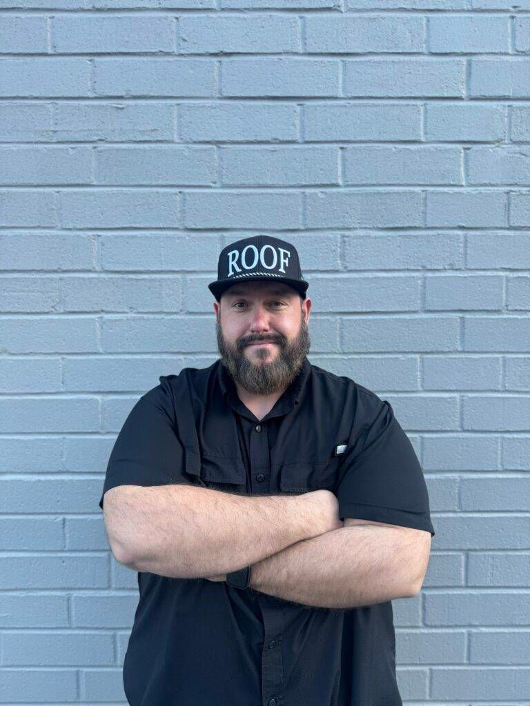 Man in black shirt and hat with "ROOF" on it, arms crossed in front of a grey brick wall.