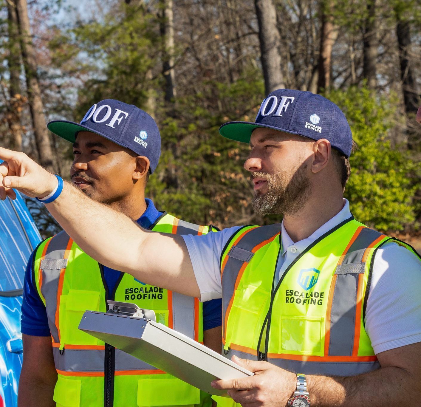 Two men in safety vests surveying a site, one pointing. Forest background.