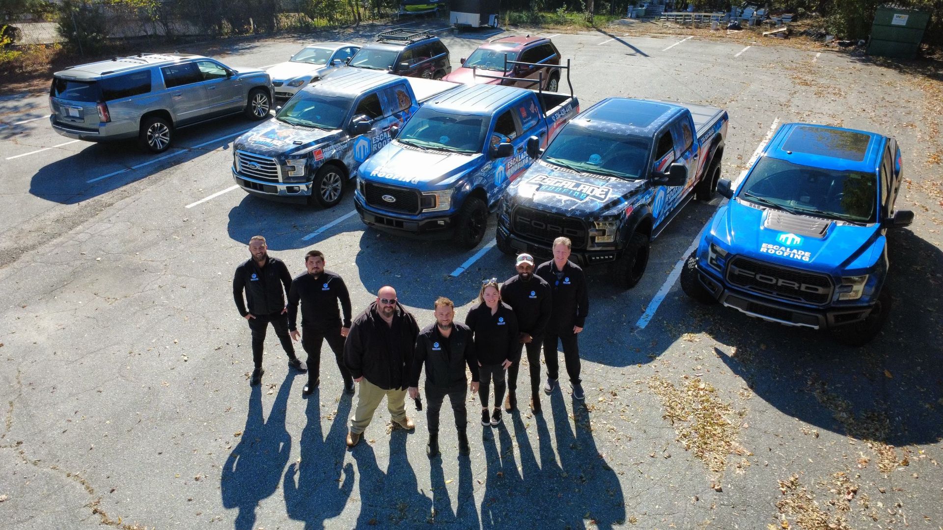 A group of people pose with multiple blue and black trucks in a parking lot.
