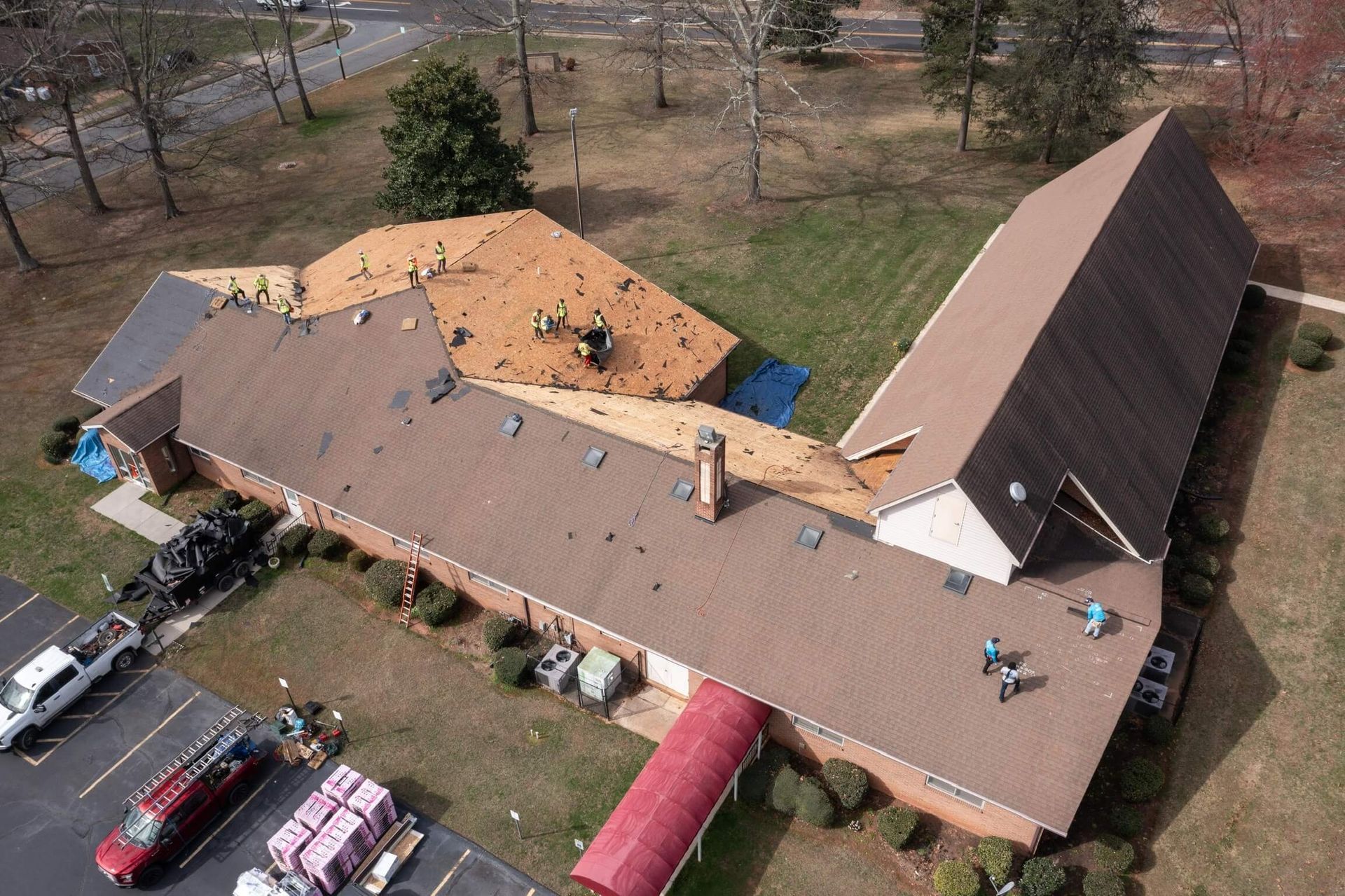 Aerial view of a building with roof repair underway. Workers on the roof, brown shingles, and parked vehicles.