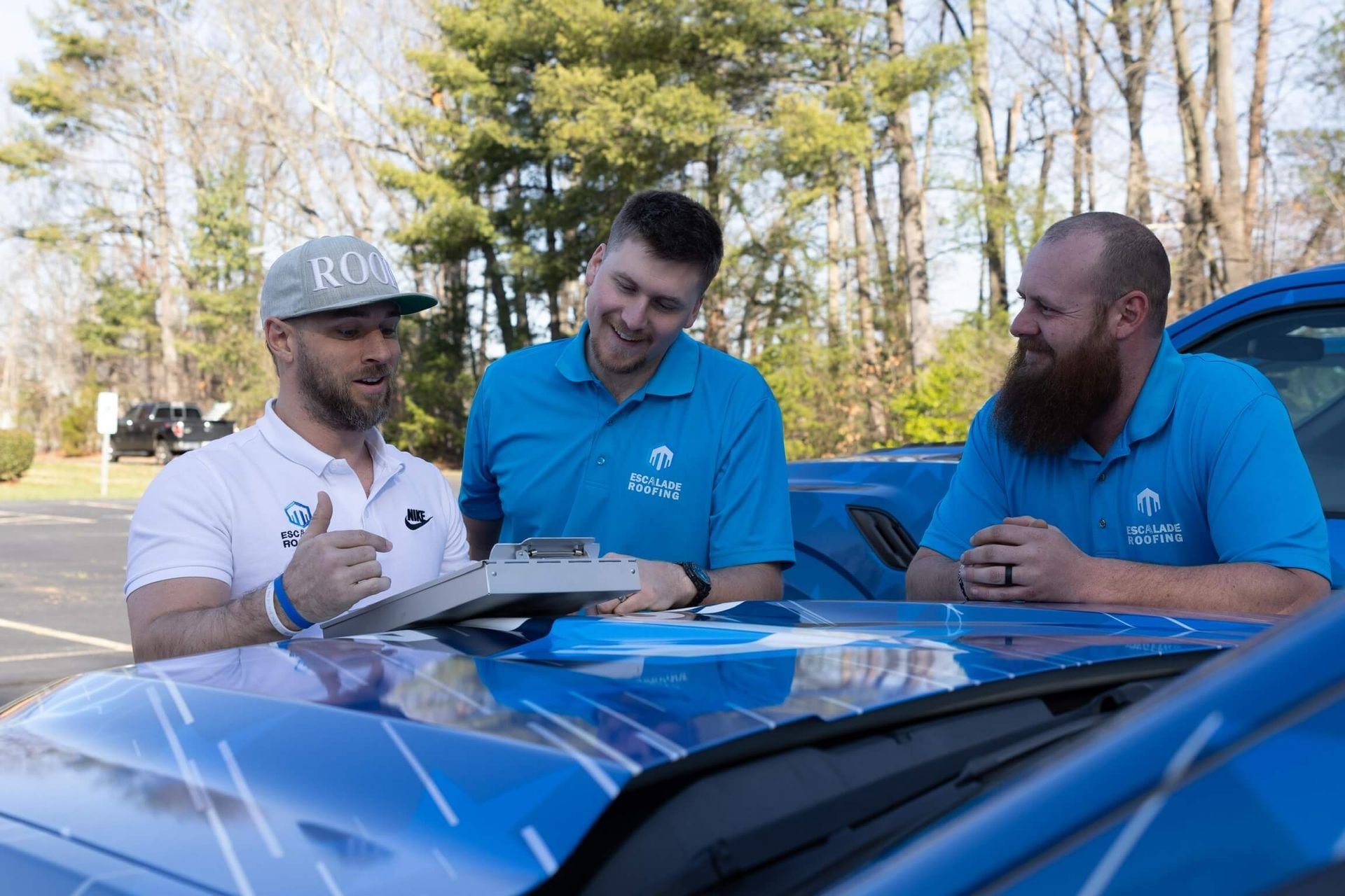 Two roofers on a roof, flexing with tools, wearing blue shirts, belts, and caps. Sunny day.