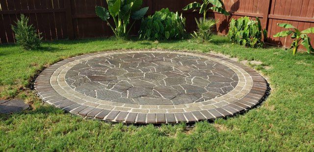 Circular stone patio in a grassy yard, surrounded by a brick border, fence and foliage in the background.