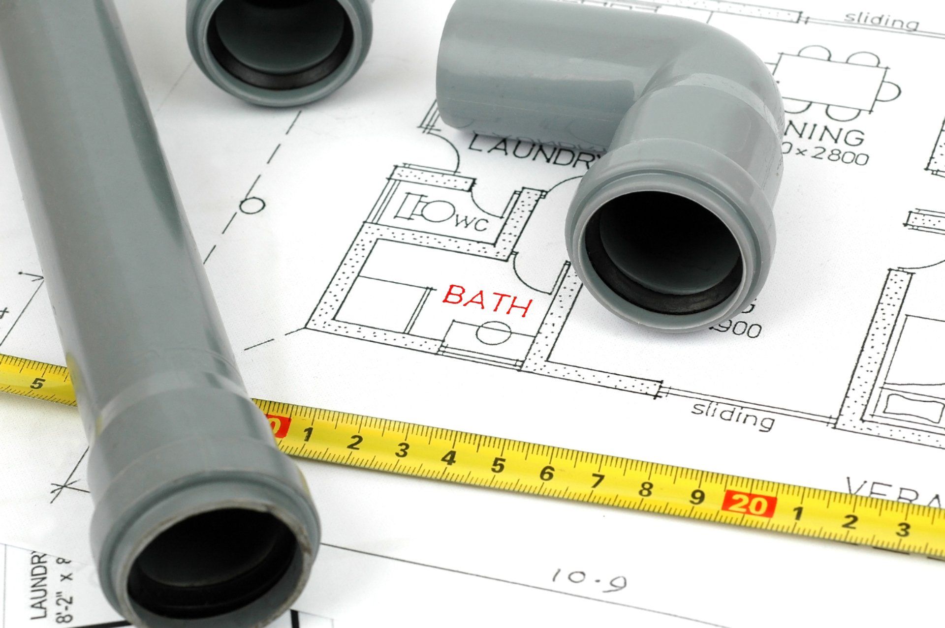 Gray plumbing pipes and elbow on a blueprint with a measuring tape, over a bathroom plan.