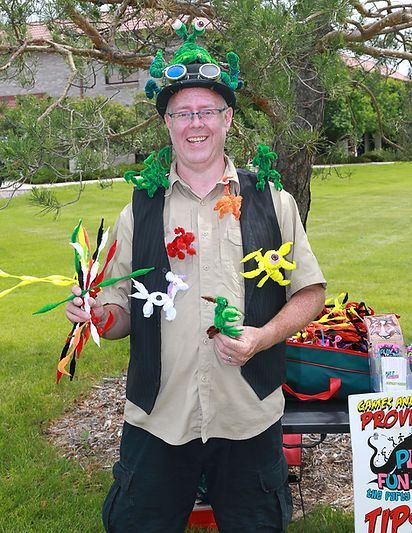 A man wearing a hat and vest is holding balloons and pipe cleaners.