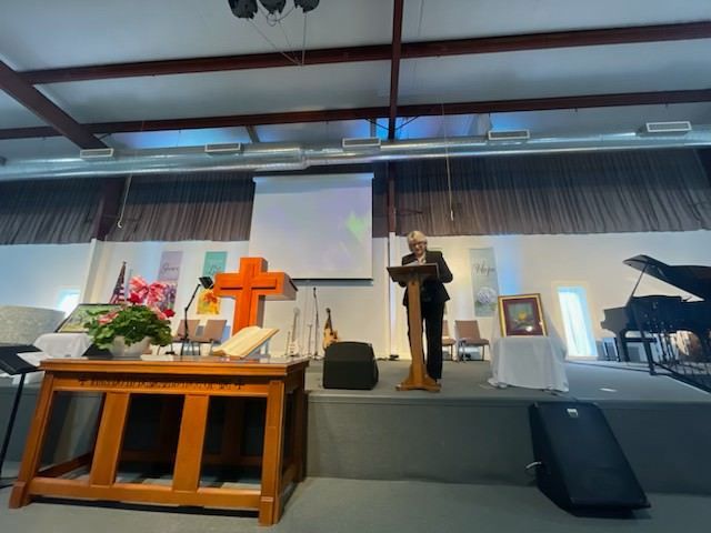 A Man Stands At A Podium In A Church With A Cross In The Background — Charleston, SC — Donna Jarrell Ministries