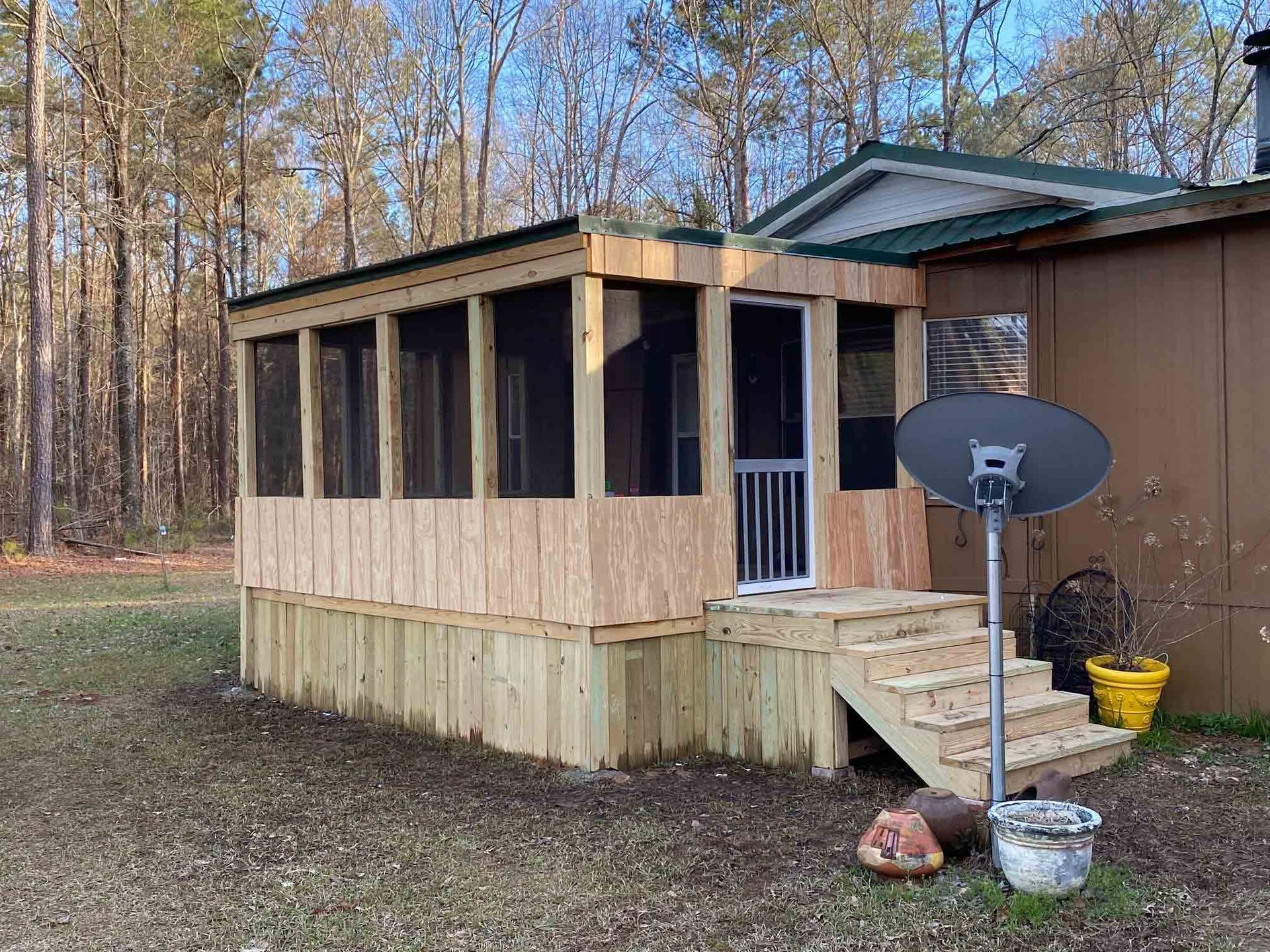 A wooden screened-in porch addition with stairs attached to the side of a house, situated in a wooded area.