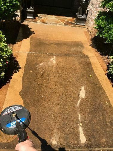 A person uses a circular surface cleaner to power wash a dirty concrete walkway leading to a house entrance.
