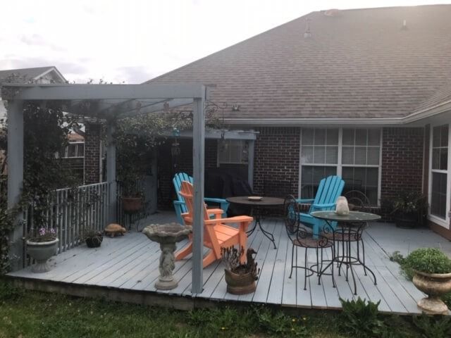 A gray backyard deck featuring a pergola, two orange Adirondack chairs, a blue chair, and small metal patio tables.