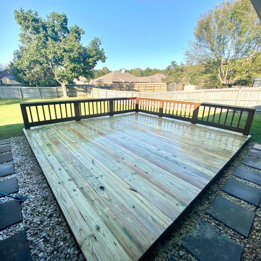 A newly built, wet wooden deck with dark railings situated in a backyard with gravel, grass, and trees.