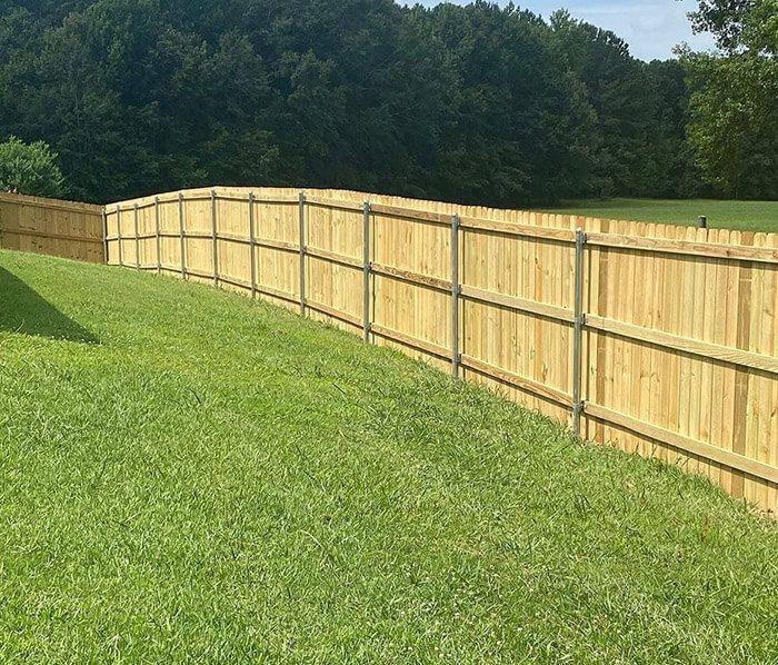 A wooden privacy fence borders a grassy, sloping yard with a background of dense green trees.