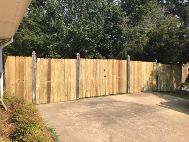 A tall, wooden privacy fence with gate panels stands on a concrete driveway in front of a tree line.