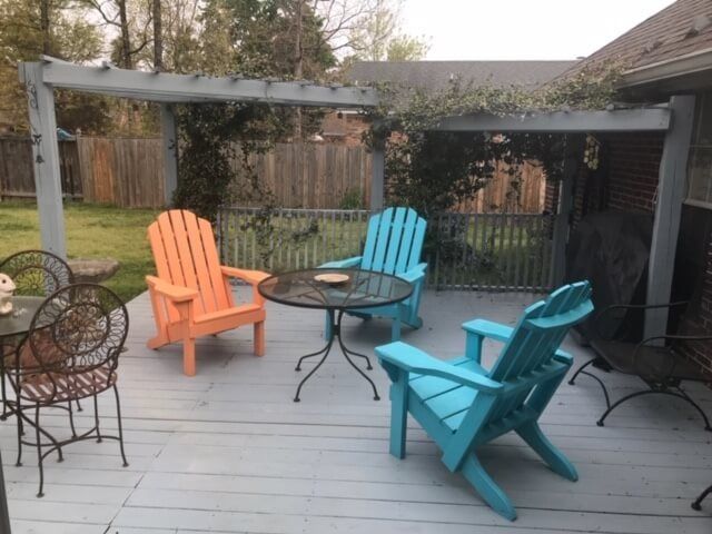 A wooden deck featuring two turquoise and one orange Adirondack chairs around a glass patio table under a pergola.