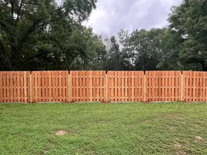A new wooden privacy fence with vertical pickets stands in a grassy backyard against a background of trees.