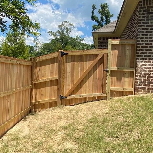 A wooden fence and gate installed on a sloped grassy area next to a brick house.