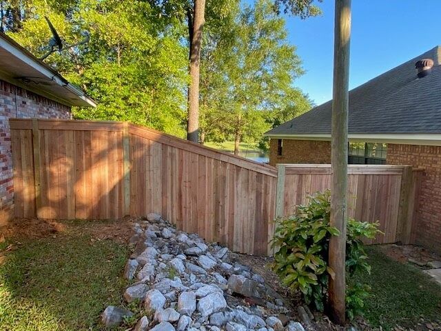 A new cedar wood privacy fence runs between two brick houses, with a stone-filled drainage ditch in the foreground.