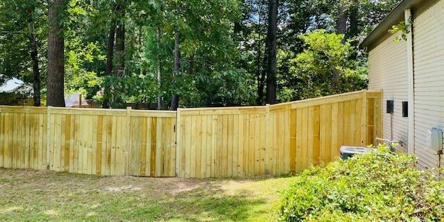 A newly installed light wood privacy fence stands in a grassy backyard next to the side of a house.