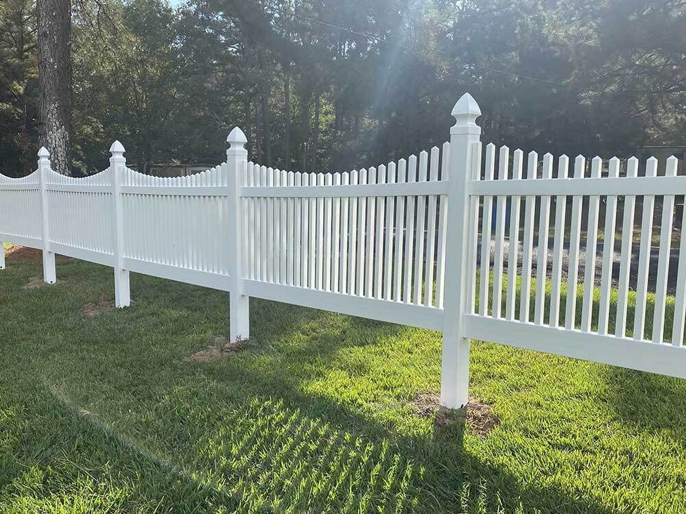 A white vinyl scalloped picket fence runs across a green grassy yard with trees in the background.