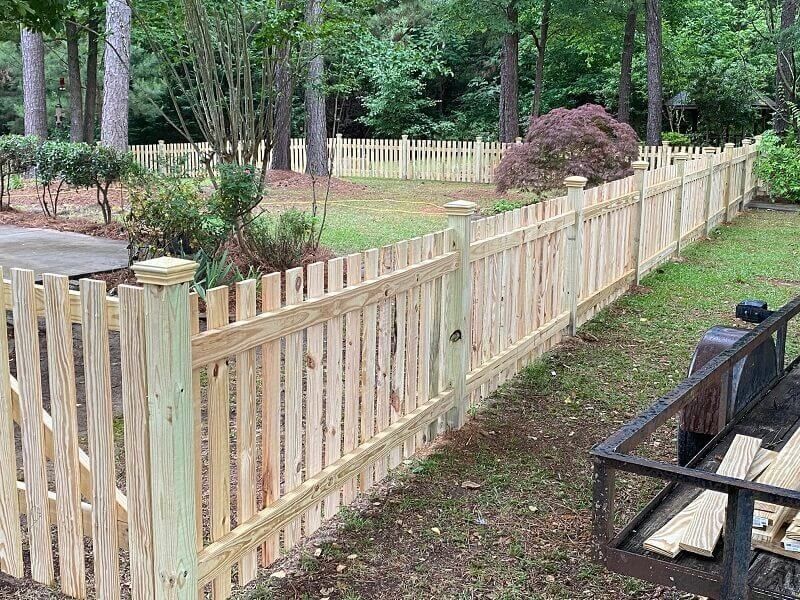 A new, light-colored wooden picket fence borders a grassy yard with trees and a small bush in the background.