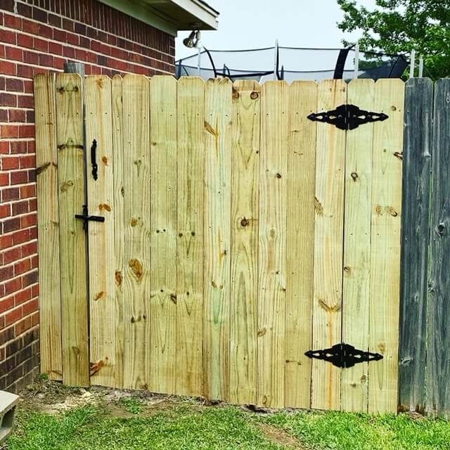A wooden gate with black ornamental hinges and a latch, set between a brick wall and an older wooden fence.
