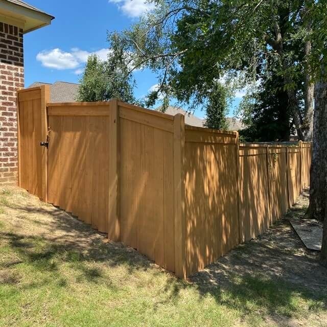 A newly installed wooden privacy fence extends from the corner of a brick house across a sloping, grassy yard.