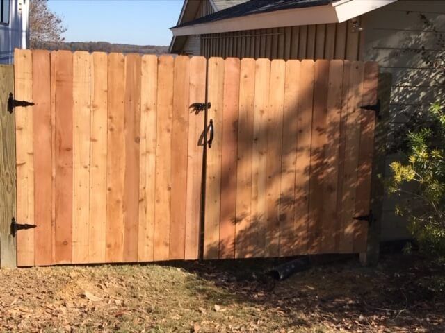 A wooden double gate stands closed against a house wall, featuring black hinges and a latch on a dirt-covered yard.