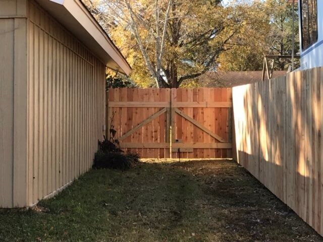 A narrow backyard area with a tan-colored wall on the left, a new wooden fence on the right, and a double-swing wood gate.
