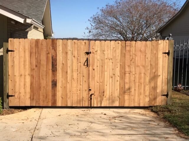 A tall wooden double gate with metal hinges sits across a concrete driveway between two residential houses.