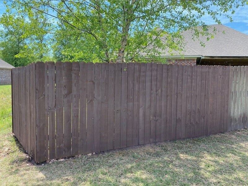 A dark brown wooden privacy fence stands in a grassy yard, partially shaded by a large green tree under a blue sky.