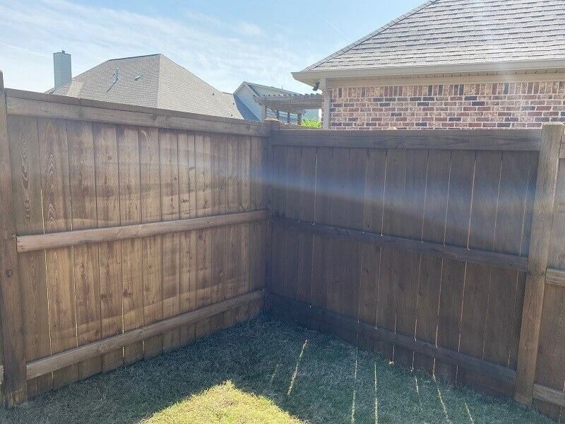 Two stained wooden fence panels meet at a corner, with brick houses visible in the background against a blue sky.