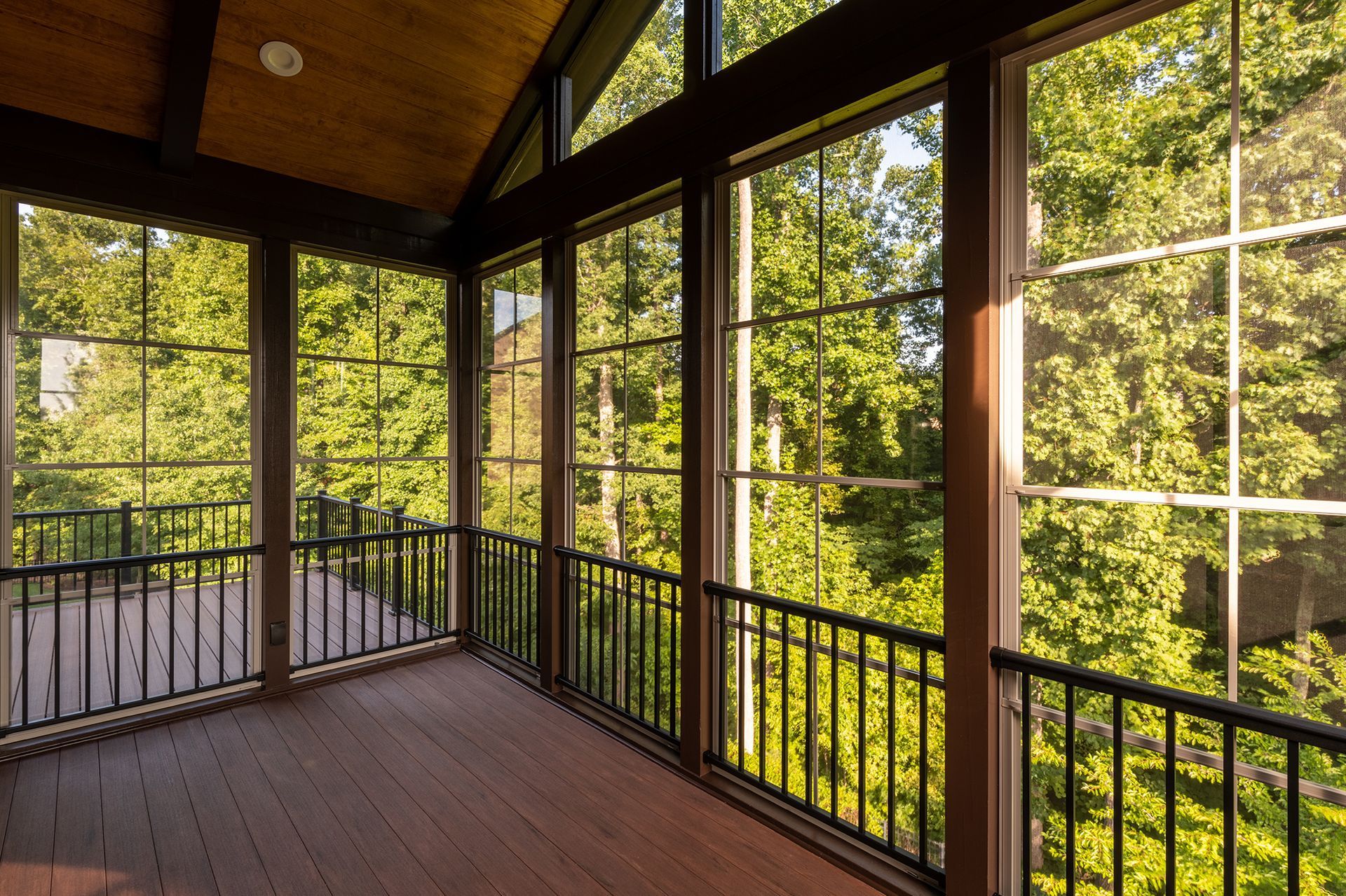 A screened-in porch with wooden decking and dark railings, looking out onto a lush green forest through large windows.