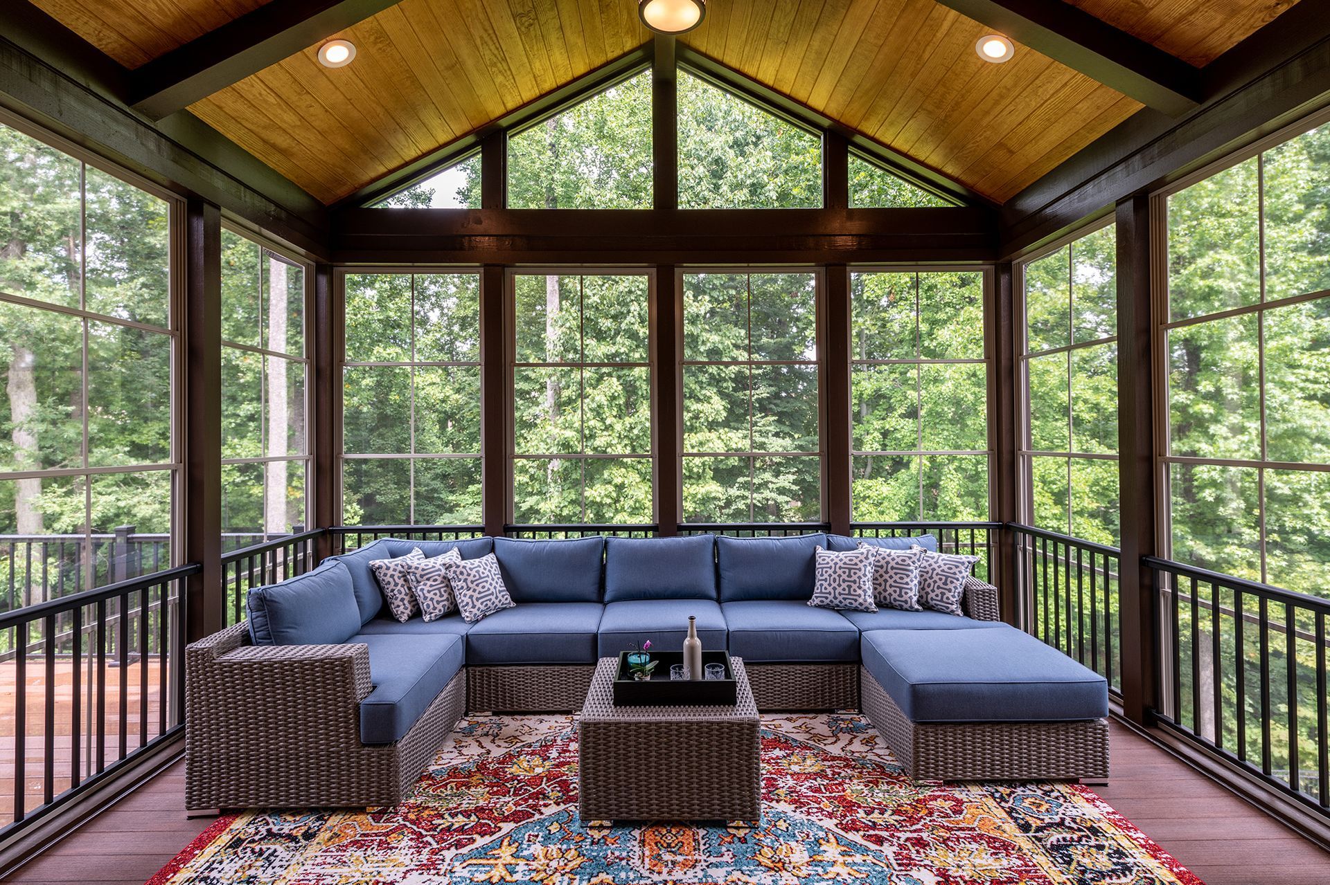 A blue sectional sofa with patterned pillows sits on a colorful rug inside a screened-in porch overlooking a forest.
