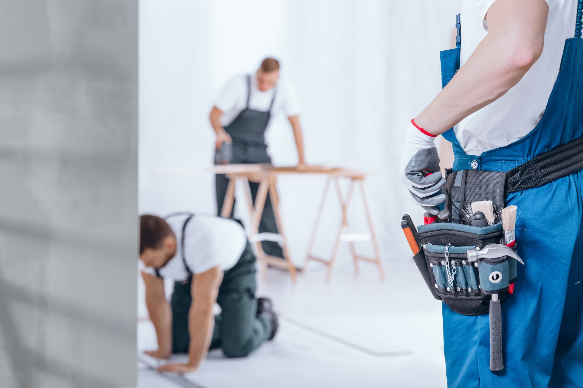 Three construction workers in blue overalls working on an interior renovation, one wearing a tool belt in the foreground.