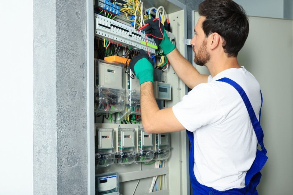 Electrician in blue overalls and green gloves working inside an electrical panel.